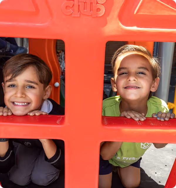 Dos niños sonrientes asomándose a través de las ventanas de un club infantil de juego rojo.