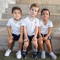 Tres niños pequeños sentados en escaleras al aire libre, vestidos con camisetas blancas y pantalones cortos oscuros.