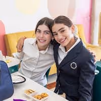Dos niñas sonrientes sentadas juntas en una mesa con comida, abrazándose amigablemente.