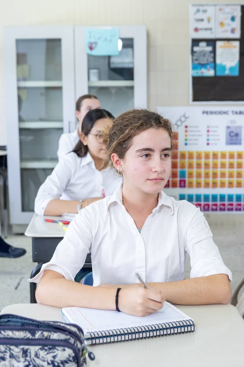Una estudiante con camisa blanca está sentada en el pupitre de una clase, con un bolígrafo en la mano y la mirada fija hacia delante. Otros alumnos están sentados detrás de ella y en la pared se ven carteles de ciencias.