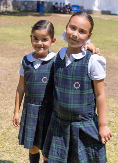 Dos niñas estudiantes sonrientes con uniforme azul a cuadros y camisa blanca, posando al aire libre en un área verde.