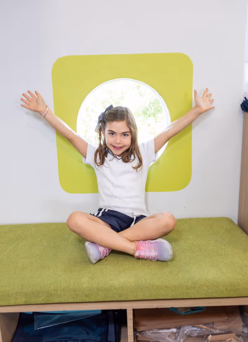 Niña sentada con las piernas cruzadas en un banco verde frente a una ventana circular con marco amarillo, sonriendo y con los brazos extendidos.