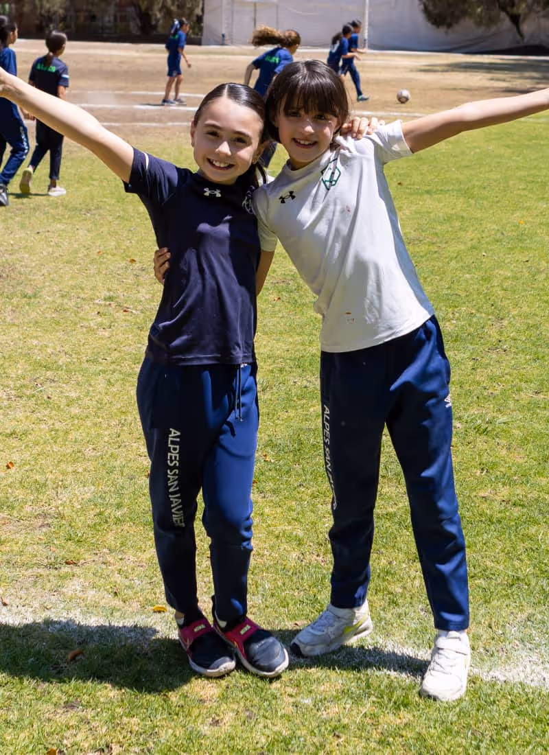 Dos niñas sonrientes con uniforme deportivo azul y blanco posan con los brazos extendidos sobre césped en un campo de deportes.