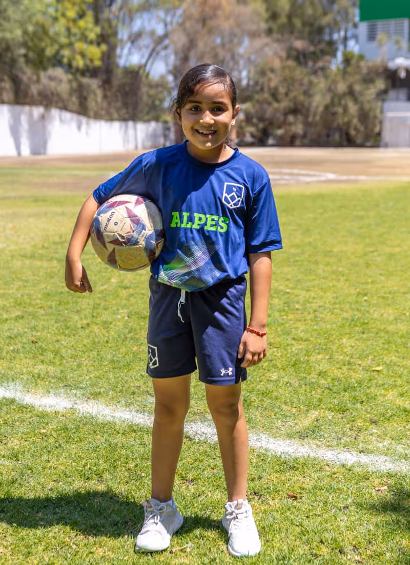 Niña sonriente con uniforme azul de fútbol que sostiene un balón en un campo verde al aire libre.
