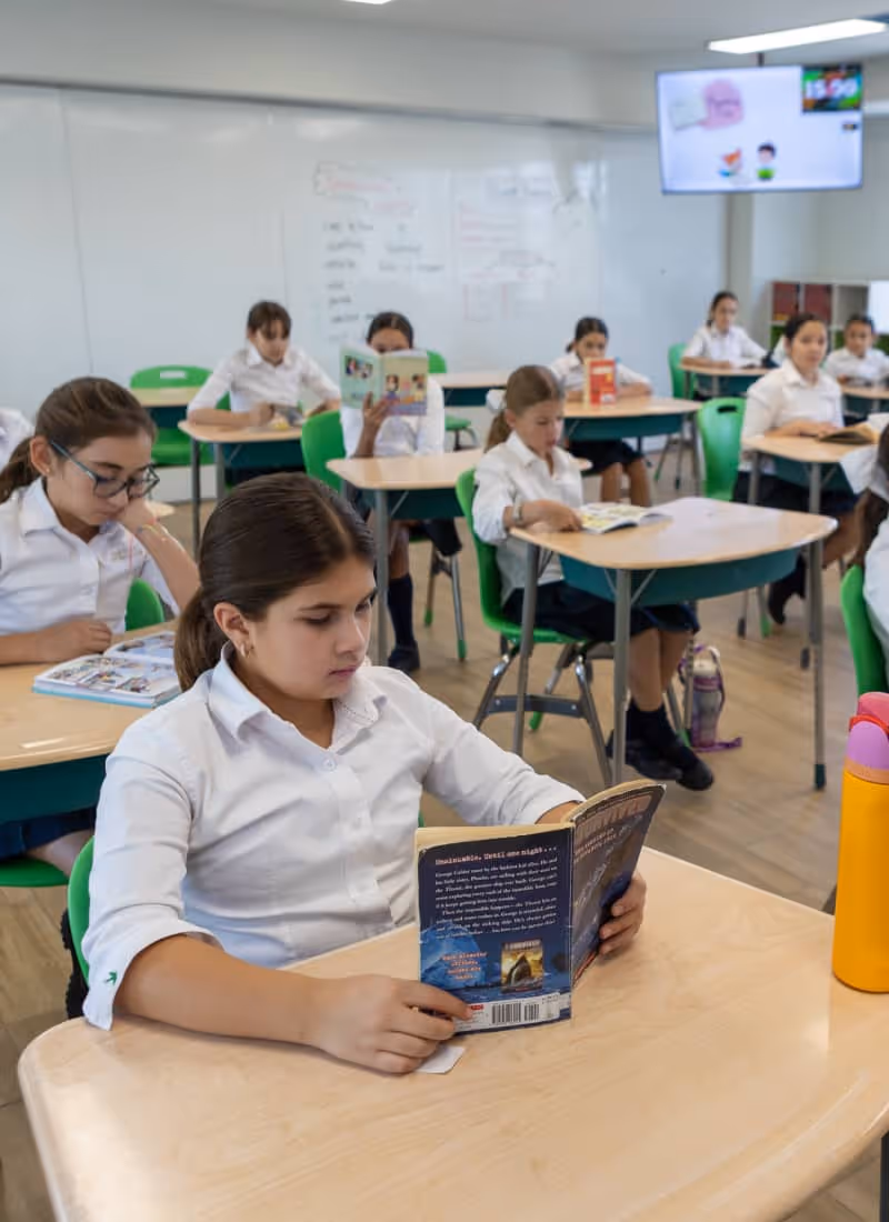 Estudiantes en uniforme blanco sentados en escritorios verdes en un salón de clases, leyendo libros.