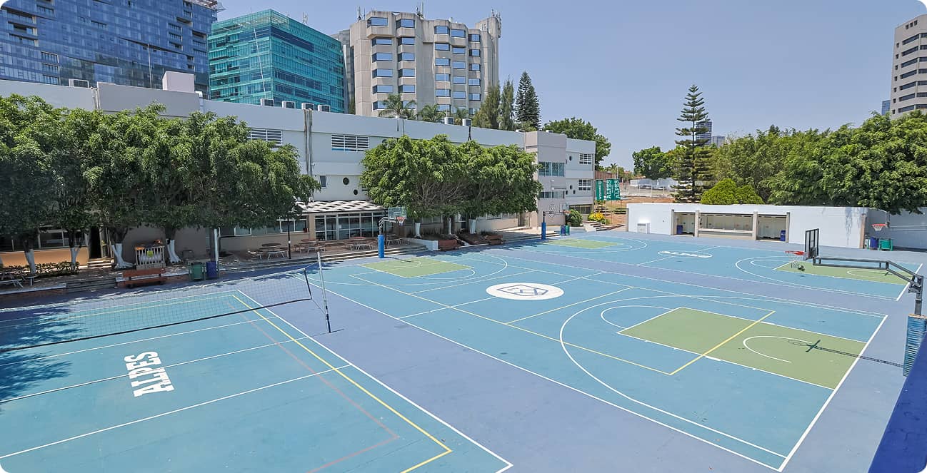 Un complejo deportivo al aire libre con canchas de baloncesto y voleibol, además de zonas para otros deportes, rodeado de árboles y adyacente a edificios modernos: un entorno ideal para programas educativos y fomentar el liderazgo.