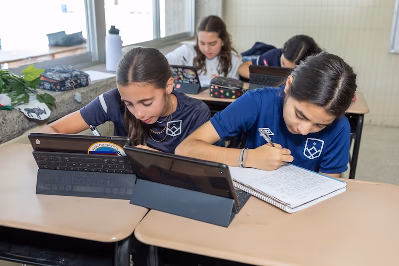 Cuatro estudiantes enfocados escribiendo en cuadernos y tabletas en un aula con escritorios.