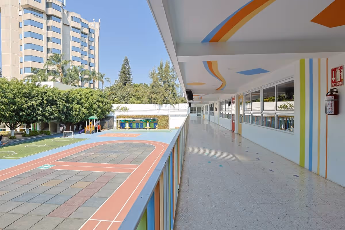 Pasillo iluminado en una escuela con ventanas, con vista a una pista deportiva y área de juegos al aire libre.