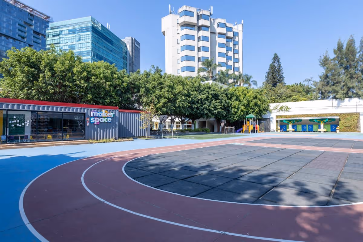 Cancha escolar al aire libre con pista de atletismo, área de juegos, árboles y edificio moderno al fondo.