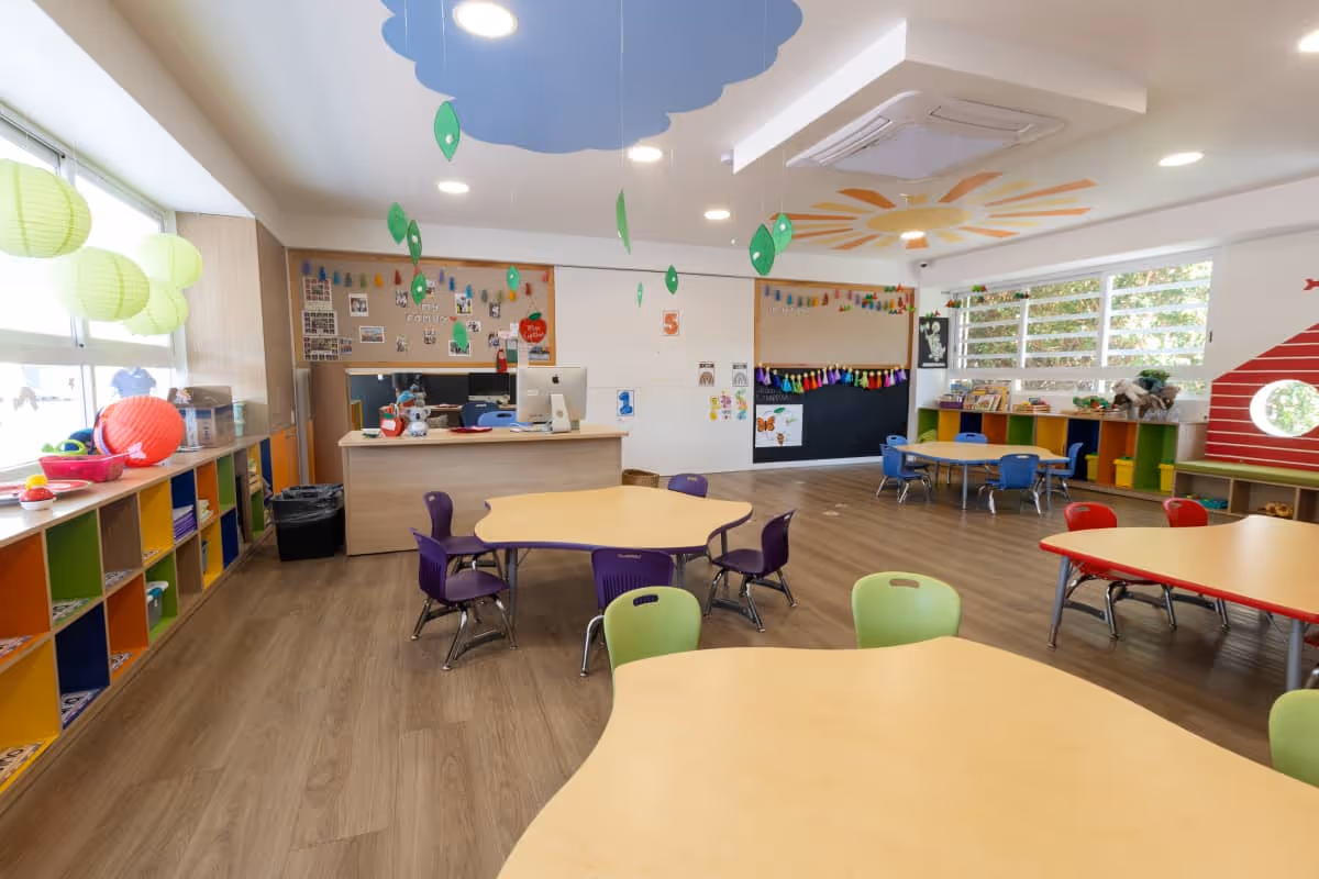 Salón de clases para niños con mesas de formas irregulares, sillas de colores, estanterías multicolor y decoraciones colgantes en el techo.