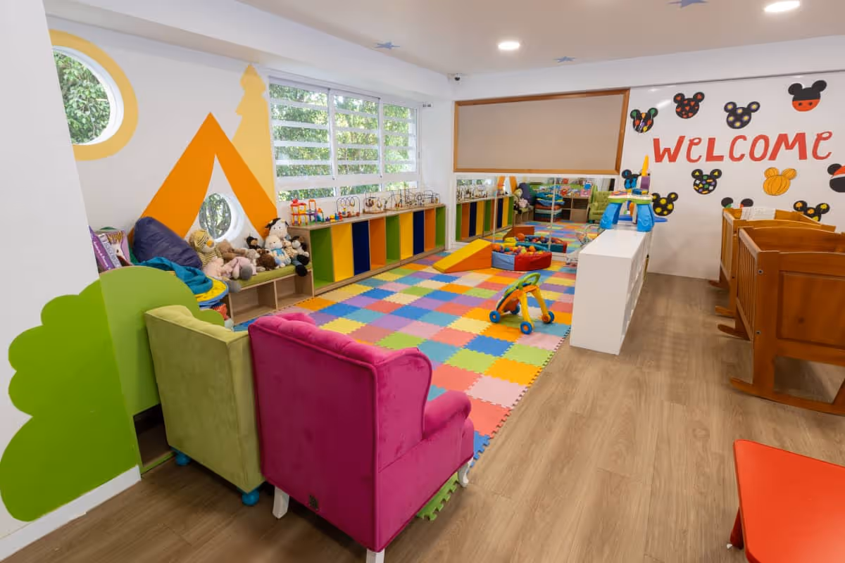 Sala de juegos para niños con piso de rompecabezas coloridos, muebles pequeños, juguetes y una pared decorada con la palabra 'Welcome' y figuras de Mickey Mouse.