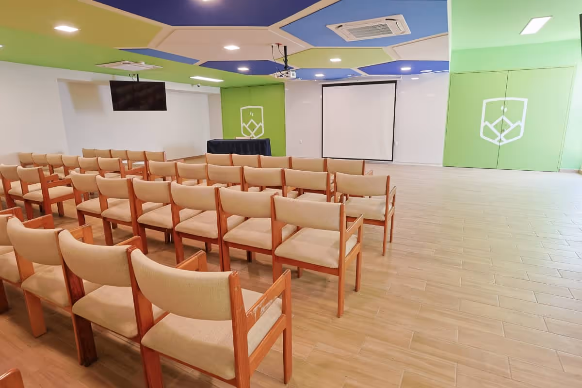 Una sala de conferencias moderna y bien iluminada, con filas de sillas acolchadas de color beige, una pantalla de proyección en la parte delantera y paredes verdes con un logotipo geométrico blanco. El techo tiene secciones azules y verdes.