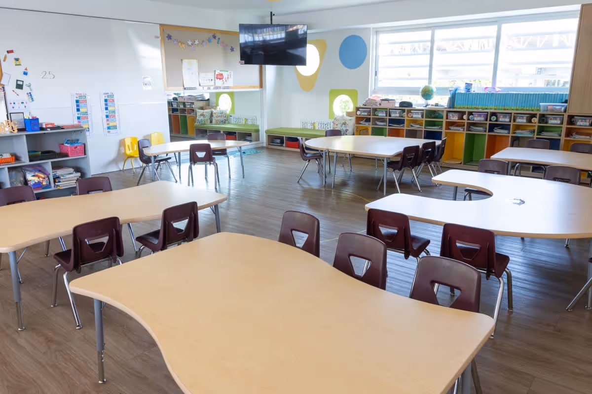 Aula escolar luminosa con sillas color marrón y mesas en forma curva, estanterías con materiales y una pantalla montada en la pared.