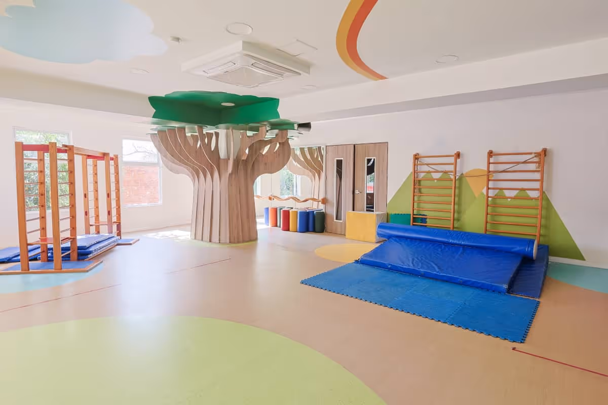Sala de juegos amplia para niños con árbol de madera decorativo, colchonetas azules y barras de escalada.