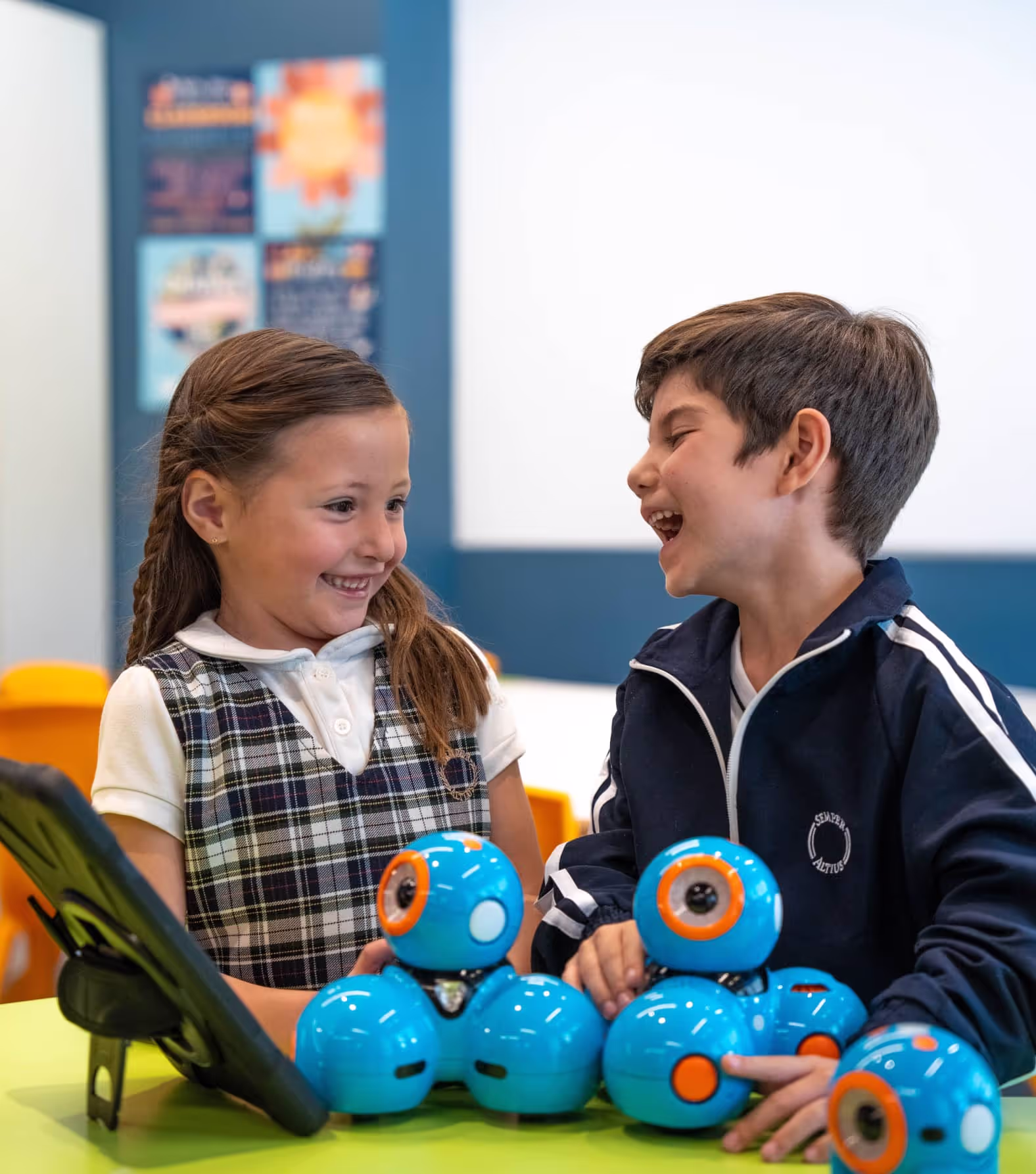 Niños sonrientes jugando con robots azules en una mesa verde dentro de un aula.