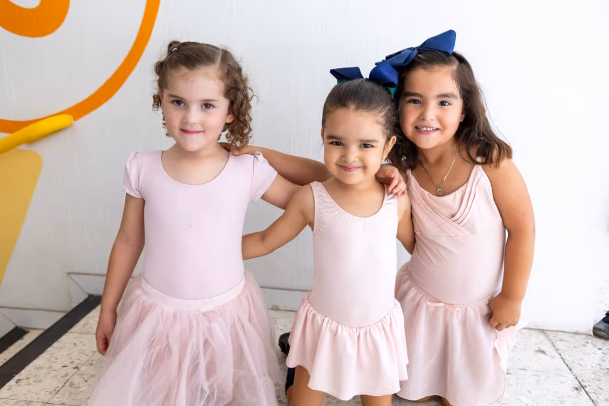 Tres niñas pequeñas vestidas con trajes rosas de ballet posando juntas y sonriendo en un espacio interior.