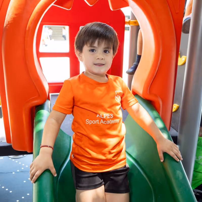 Niño con camiseta naranja de ALPES Sport Academy sentado en un tobogán verde en parque infantil.