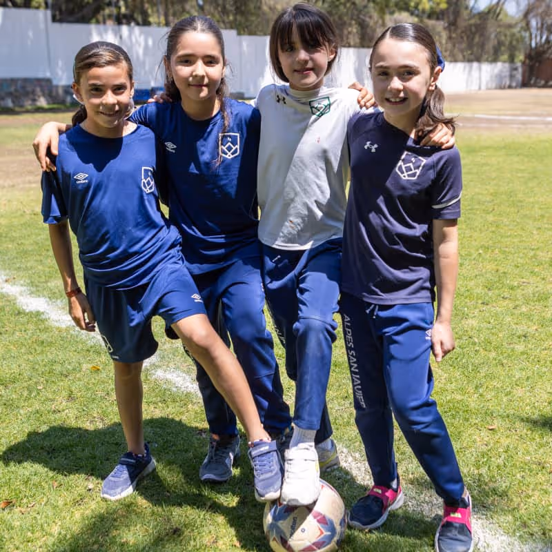 Cuatro niñas en uniforme deportivo azul posando juntas, una con el pie sobre una pelota de fútbol en un campo de césped.