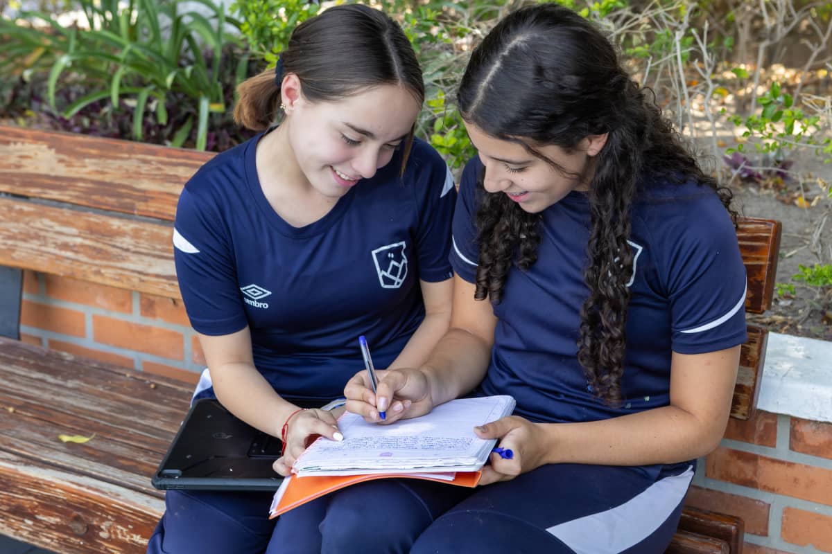 Dos estudiantes sentadas en un banco al aire libre, compartiendo y escribiendo en una libreta mientras sonríen.