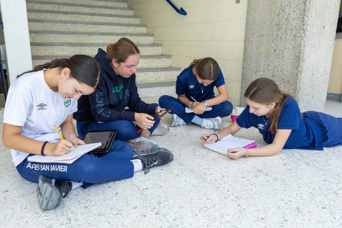 Cuatro estudiantes sentadas en el suelo junto a unas escaleras, escribiendo y usando una tableta para estudiar.