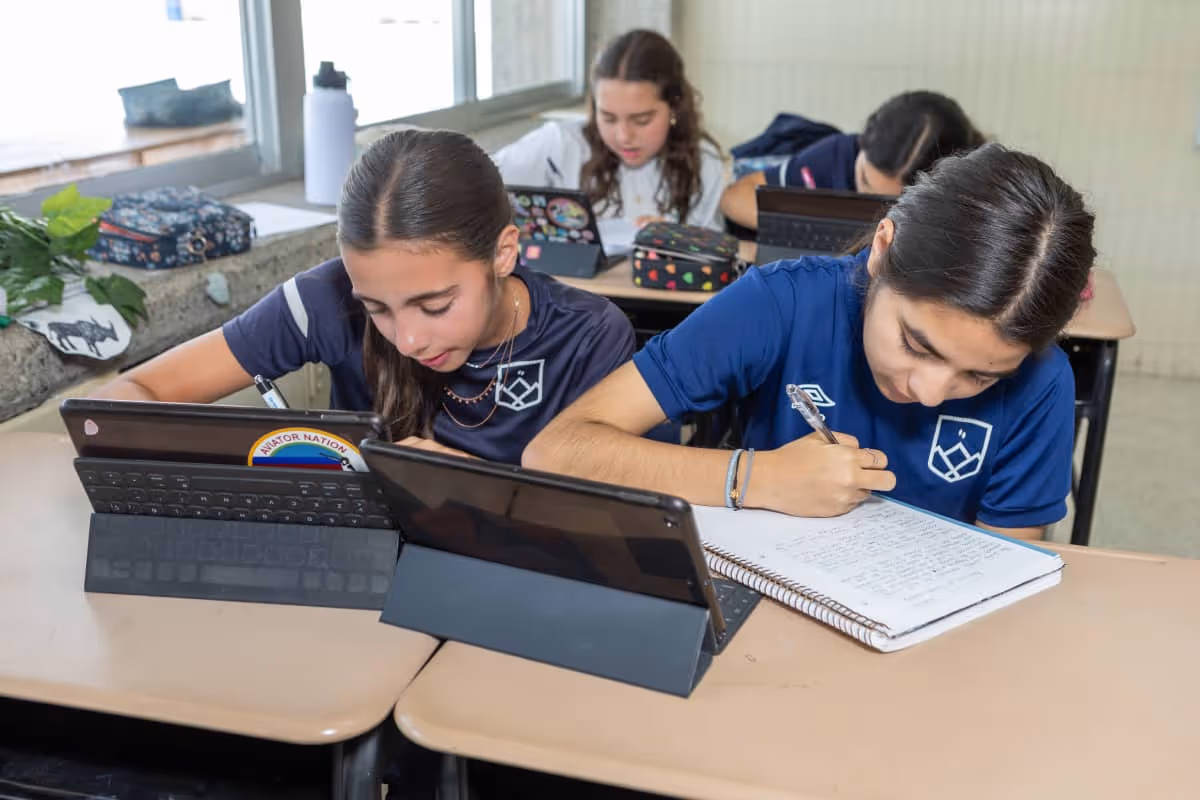 Cuatro estudiantes escriben usando tabletas y cuadernos en un salón de clases con luz natural.